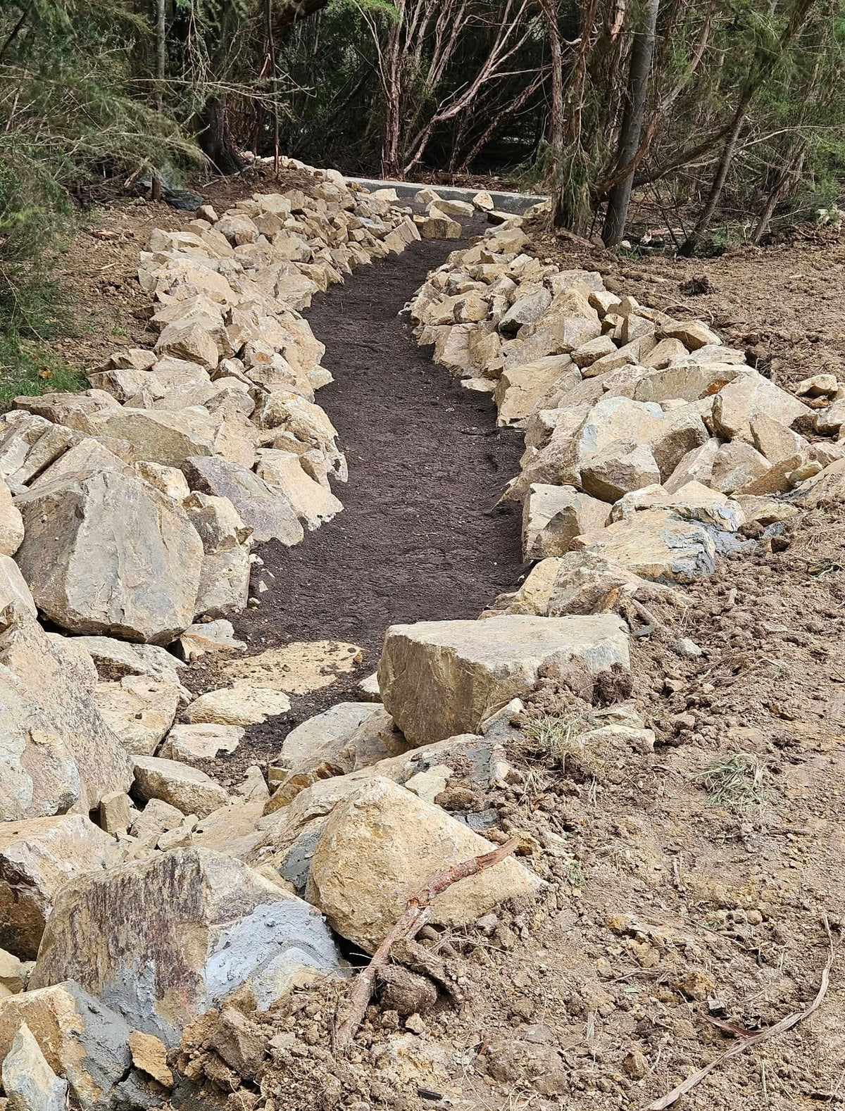 A stone-lined pathway or drainage channel with dark soil in the center, surrounded by large light-colored rocks and trees in an outdoor wooded setting.