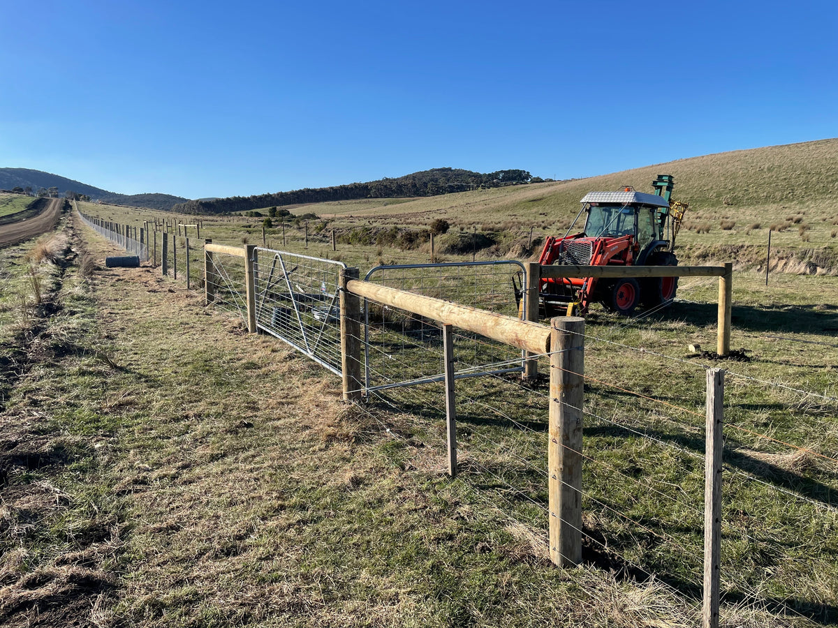 A wooden and wire fence line installed in an open grassy field, with a red tractor parked nearby and hills under a clear blue sky in the background.